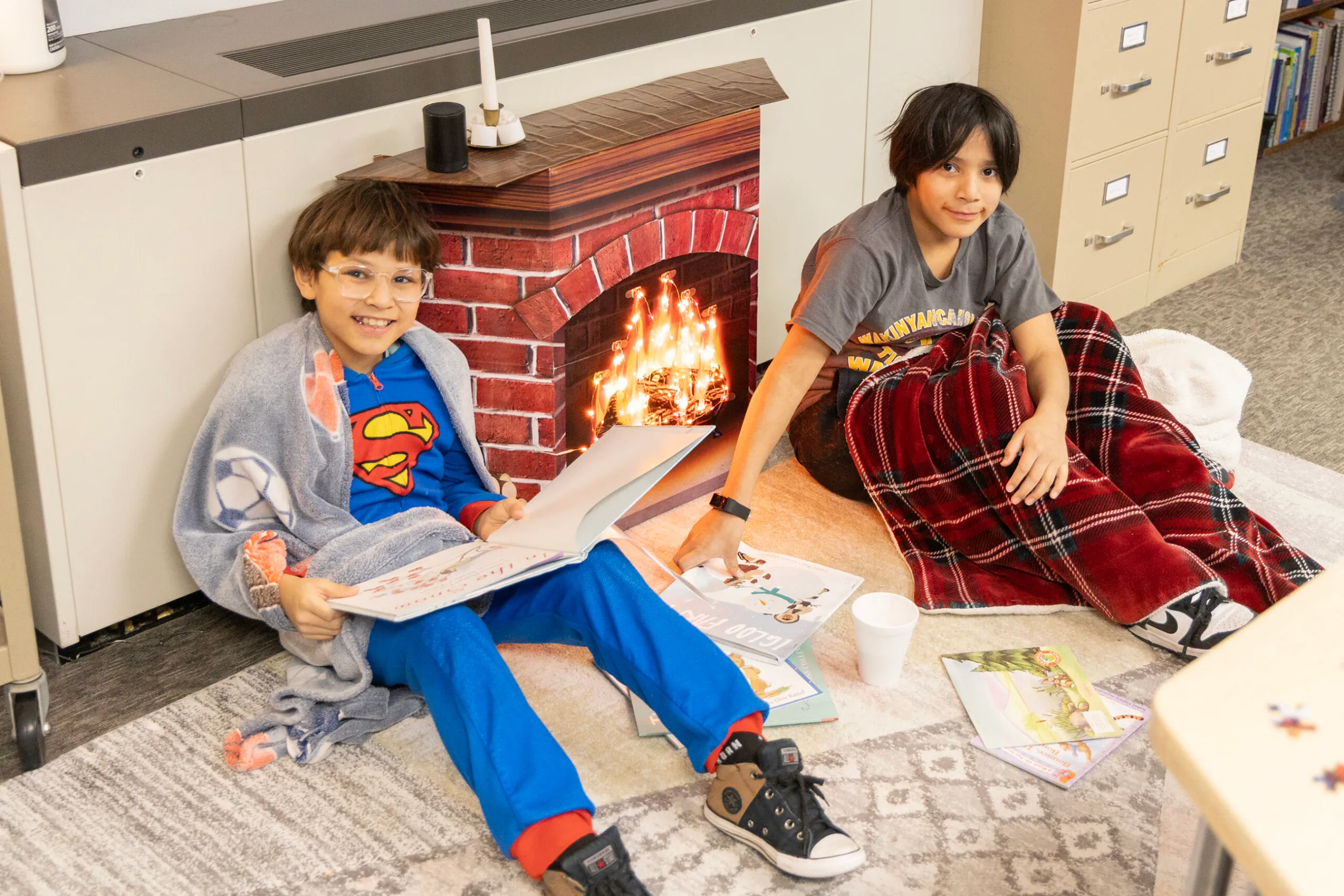 Eingehüllt in Decken machen es sich zwei Jungen am "Snugs & Hugs Day" an einem imaginären Lagerfeuer gemütlich, um zu lesen und warmen Kakao zu trinken.